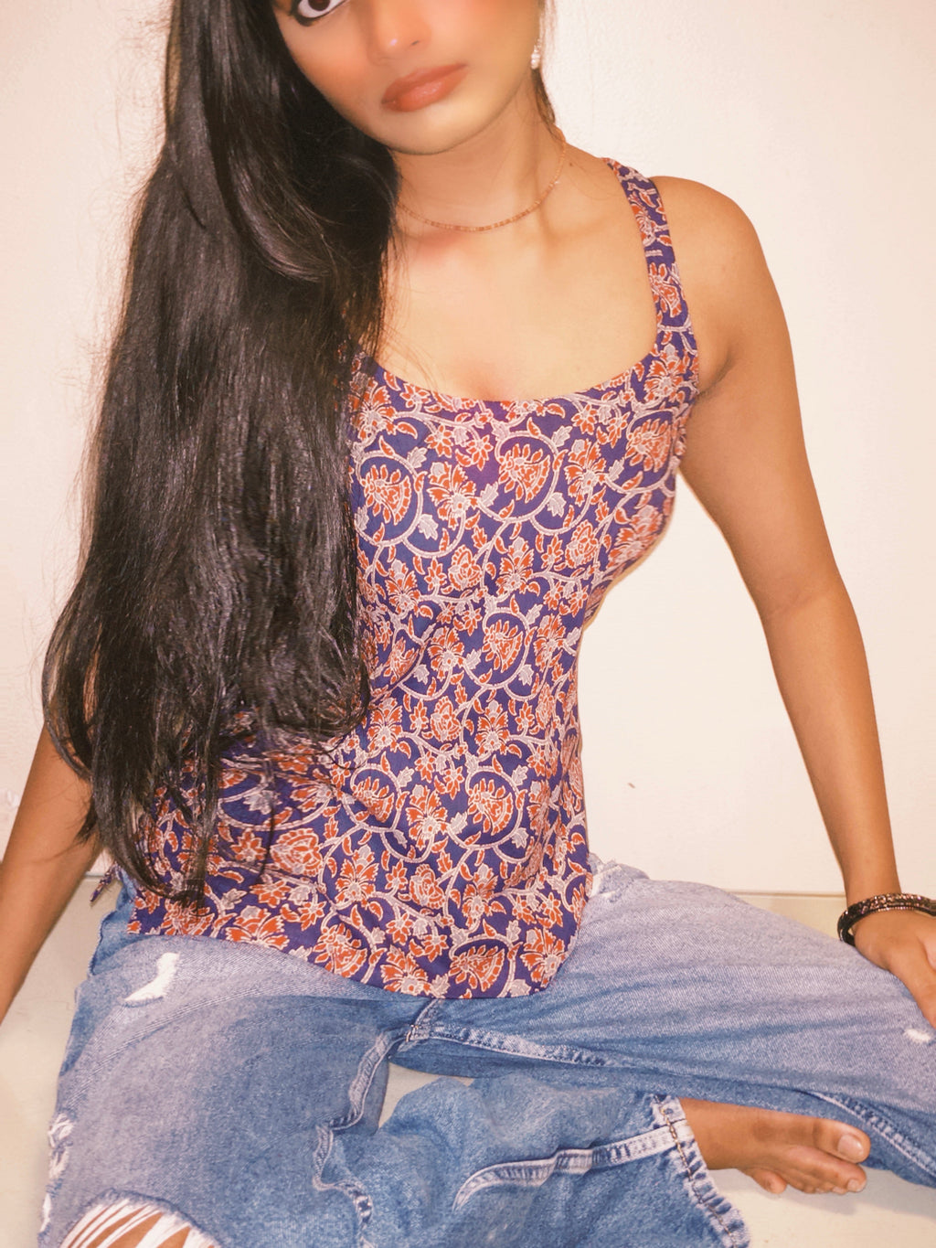 Person wearing a patterned top and jeans sitting on a couch. A woman wearing 100% cotton Deep indigo blue short kurti with white bandhani-inspired or floral motifs, halter neck with V-cut front, backless criss-cross tie-back, sleeveless, and a straight short kurti length with a gathered hem.