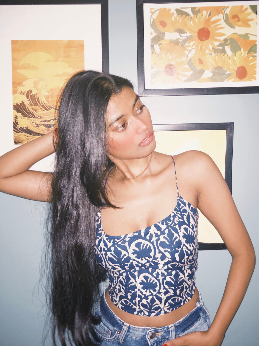 Woman wearing a blue patterned top and denim shorts standing against a wall with floral artwork. Surzaa backless blue and white patterned crop top with a lace-up back. 