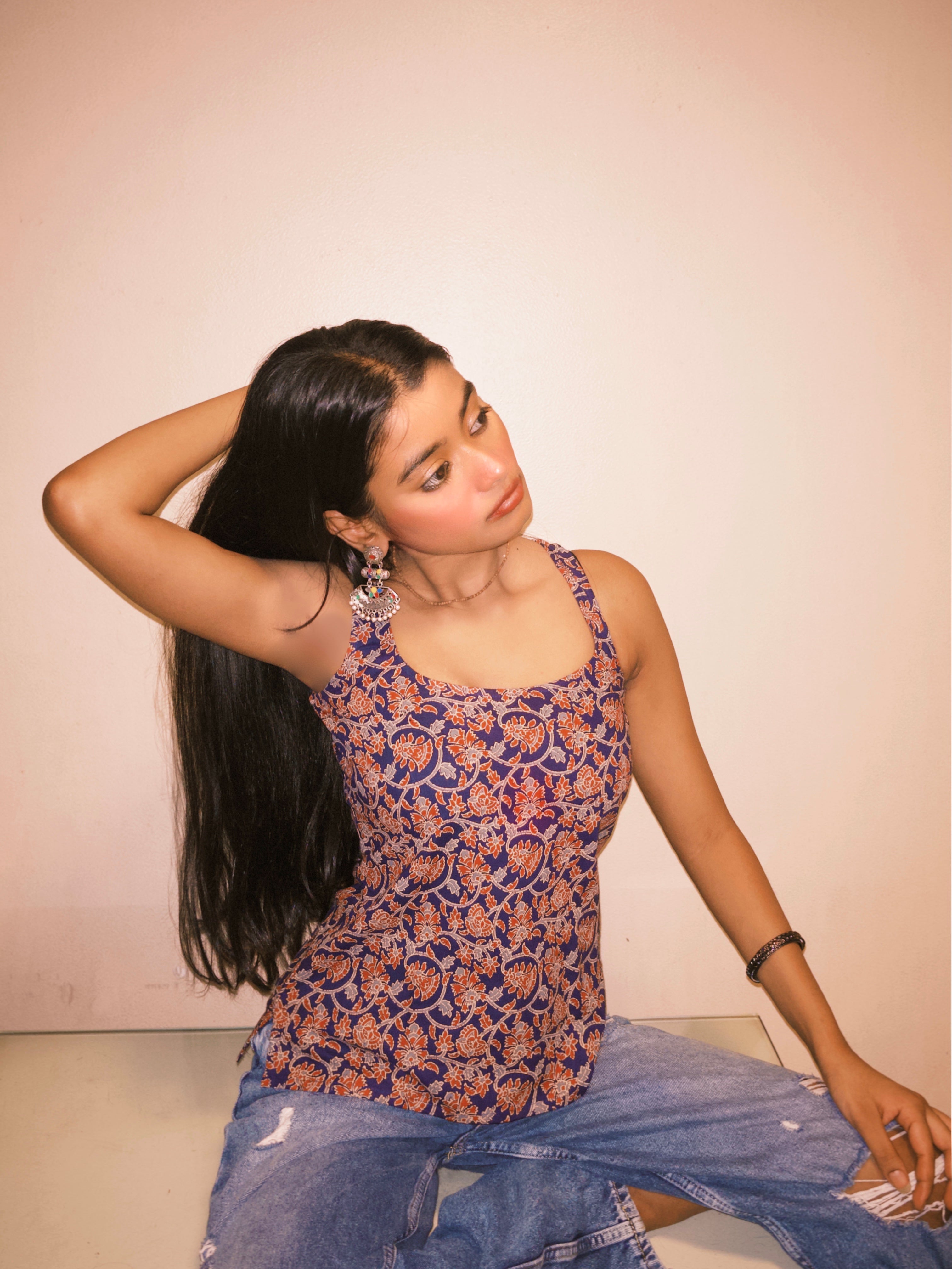 Woman wearing a patterned top and jeans sitting on a light-colored surface with a plain background. A woman wearing 100% cotton Deep indigo blue short kurti with white bandhani-inspired or floral motifs, halter neck with V-cut front, backless criss-cross tie-back, sleeveless, and a straight short kurti length with a gathered hem.