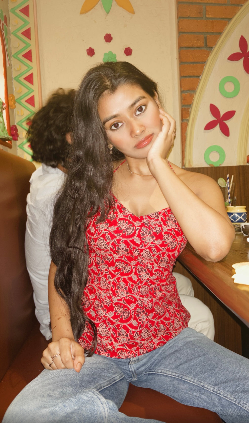 Woman in a red patterned top and jeans sitting in a casual setting. A woman modeling a red and white printed, backless, knot-back top with blue jeans, alongside product details including the name 'Rakt - Red Backless Knot-Back Printed Top'.