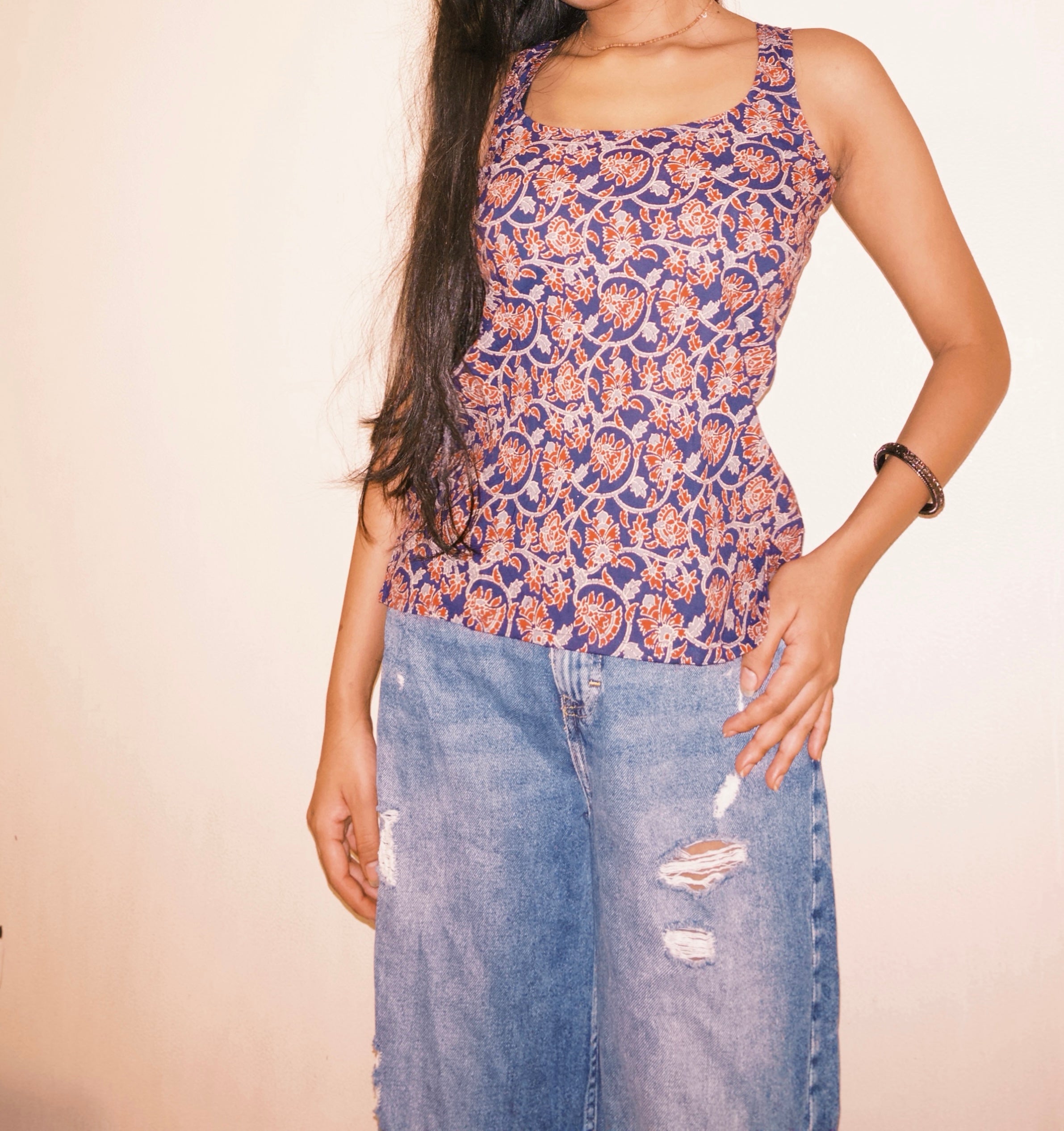 Person wearing a patterned top and blue jeans on a plain background. A woman wearing 100% cotton Deep indigo blue short kurti with white bandhani-inspired or floral motifs, halter neck with V-cut front, backless criss-cross tie-back, sleeveless, and a straight short kurti length with a gathered hem.