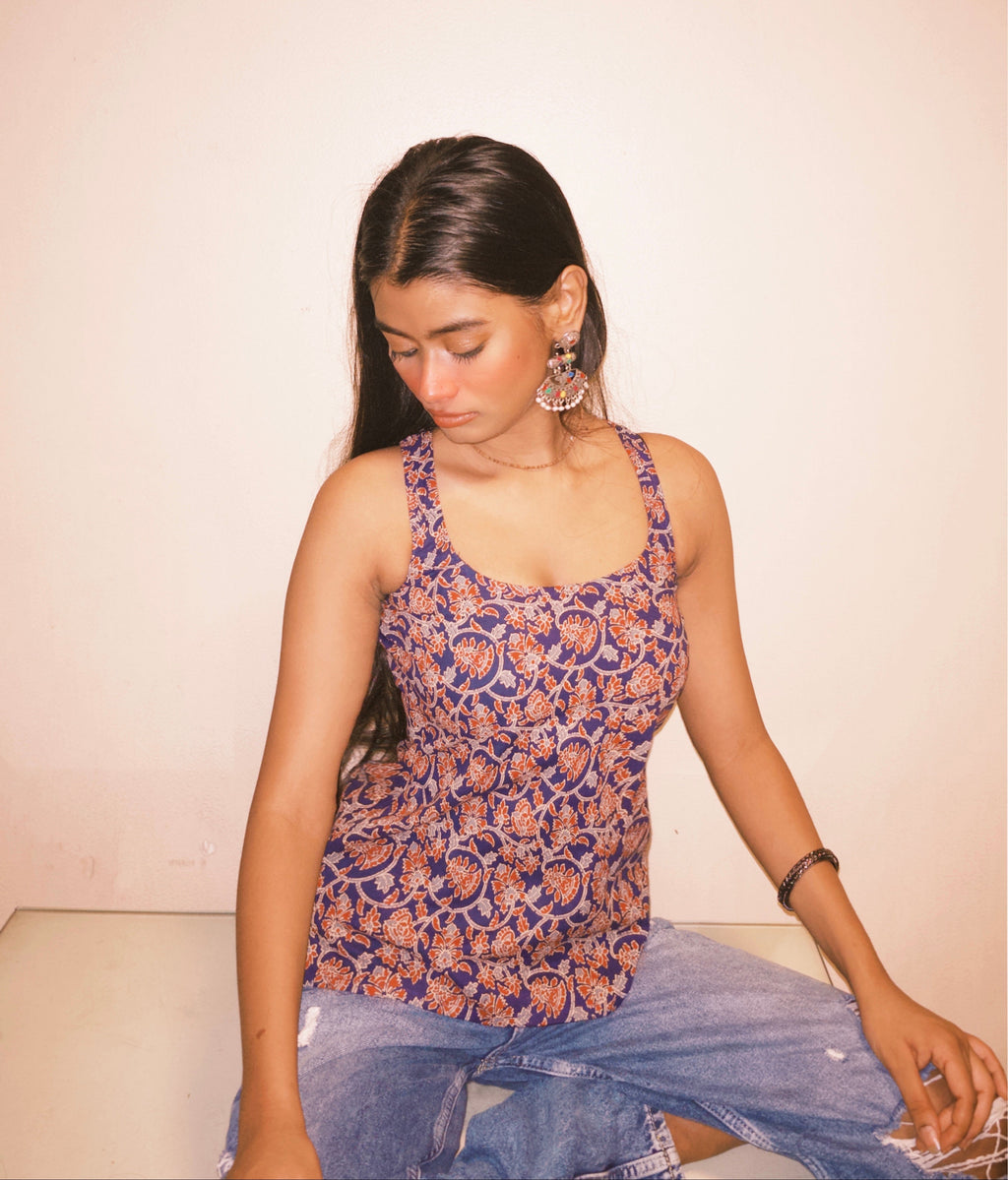 Woman wearing a patterned top and jeans sitting on a white surface with a plain background. A woman wearing 100% cotton Deep indigo blue short kurti with white bandhani-inspired or floral motifs, halter neck with V-cut front, backless criss-cross tie-back, sleeveless, and a straight short kurti length with a gathered hem.
