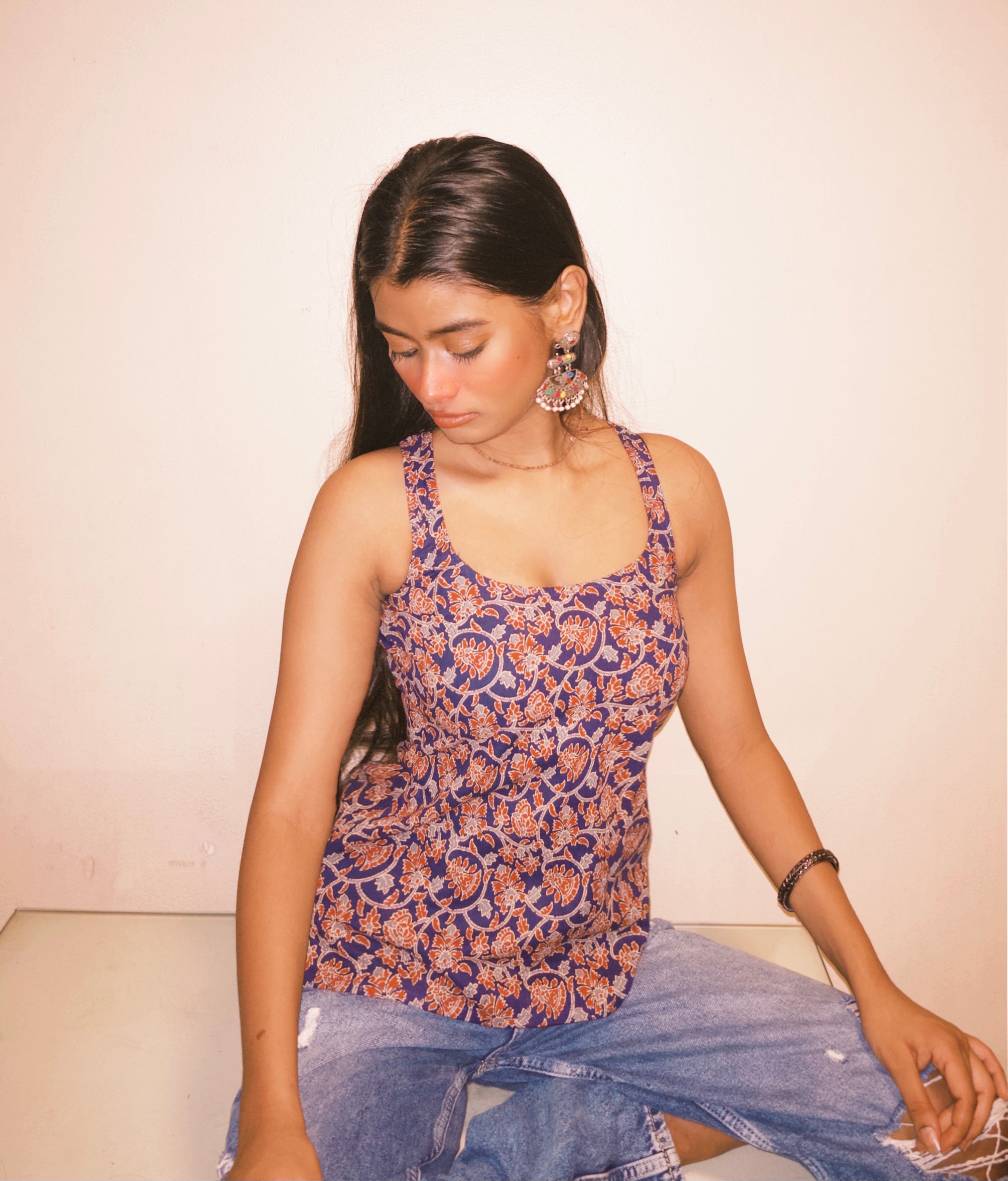 Woman wearing a patterned top and jeans sitting on a white surface with a plain background. A woman wearing 100% cotton Deep indigo blue short kurti with white bandhani-inspired or floral motifs, halter neck with V-cut front, backless criss-cross tie-back, sleeveless, and a straight short kurti length with a gathered hem.